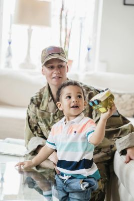 A young boy joyfully plays with a toy while his military parent watches in a cozy living room.