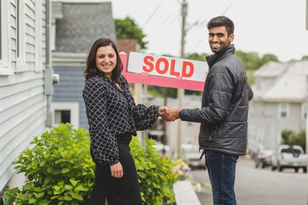 Two people shake hands in front of a sold sign, celebrating a real estate deal.