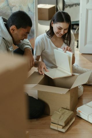 A young couple joyfully unpacks boxes in their new home, surrounded by books and cardboard packages.